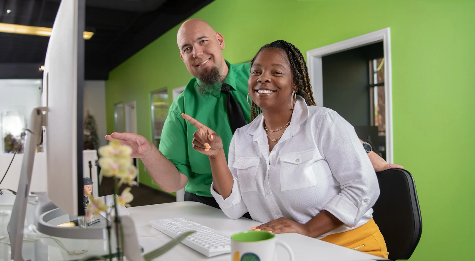 A man and woman excitedly pointing at a computer screen with bright green wall behind.